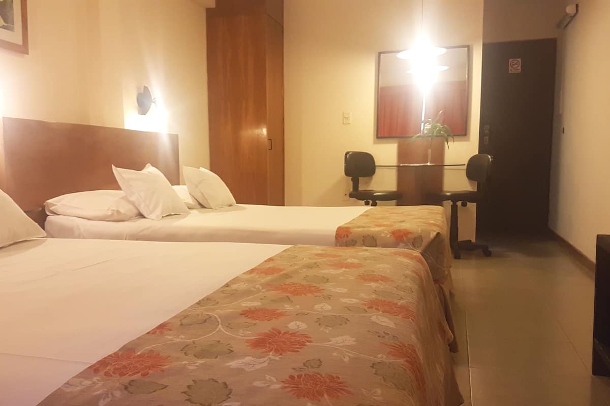 Habitación de hotel con dos camas individuales, cabecera de madera, mesita de noche, escritorio y espejo.