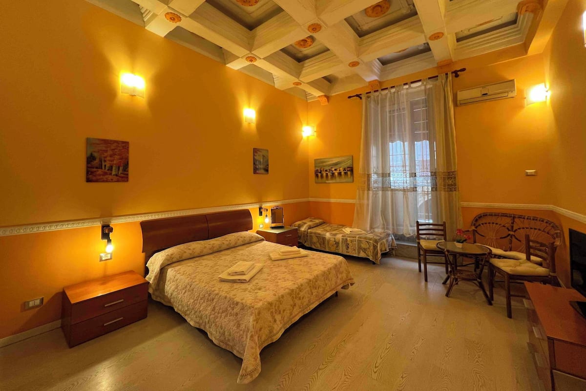Une chambre d’hôtel comprenant un lit, deux tables de chevet, une chaise, une petite table et une fenêtre avec des rideaux.