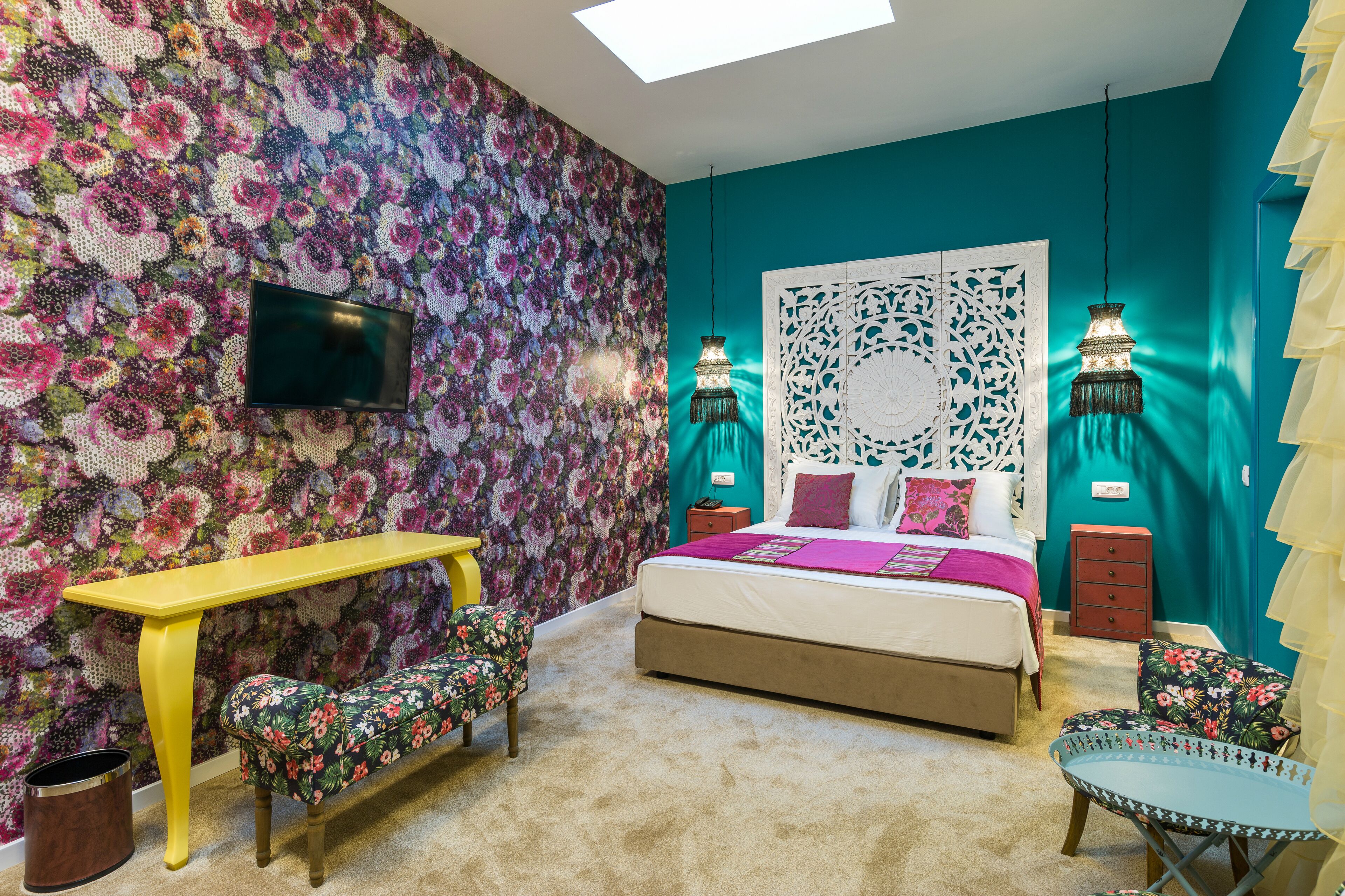 Quarto com papel de parede floral, uma cama com cabeceira branca, uma escrivaninha amarela, um banco e duas mesas de cabeceira com luminárias.