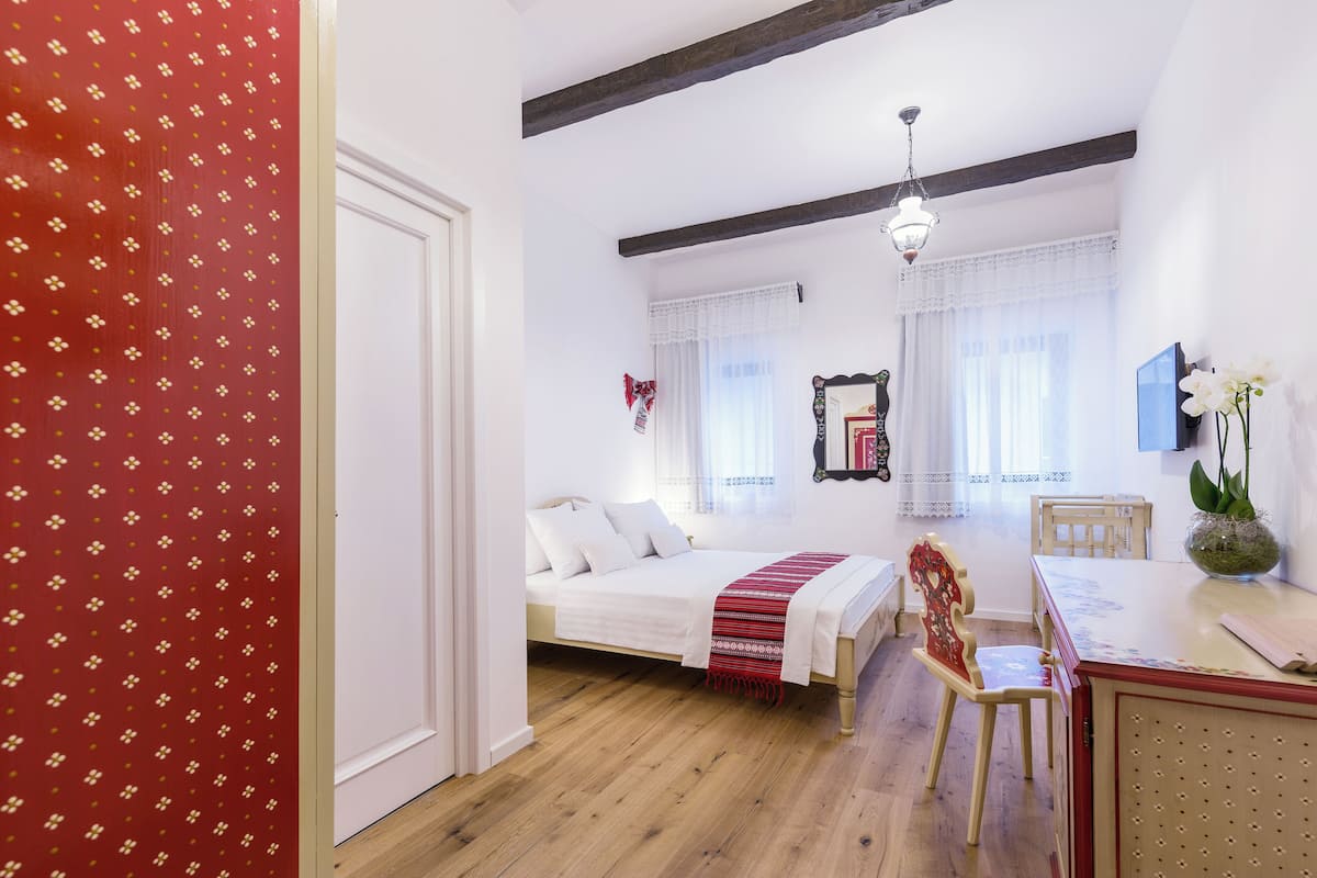 A bedroom with a bed, a wooden floor, a dining table with chairs, and a window with curtains.