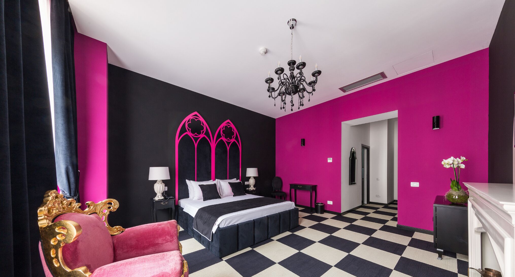 A modern bedroom with a black and white checkered floor, a bed with black and white bedding, a chandelier, and a pink accent wall.