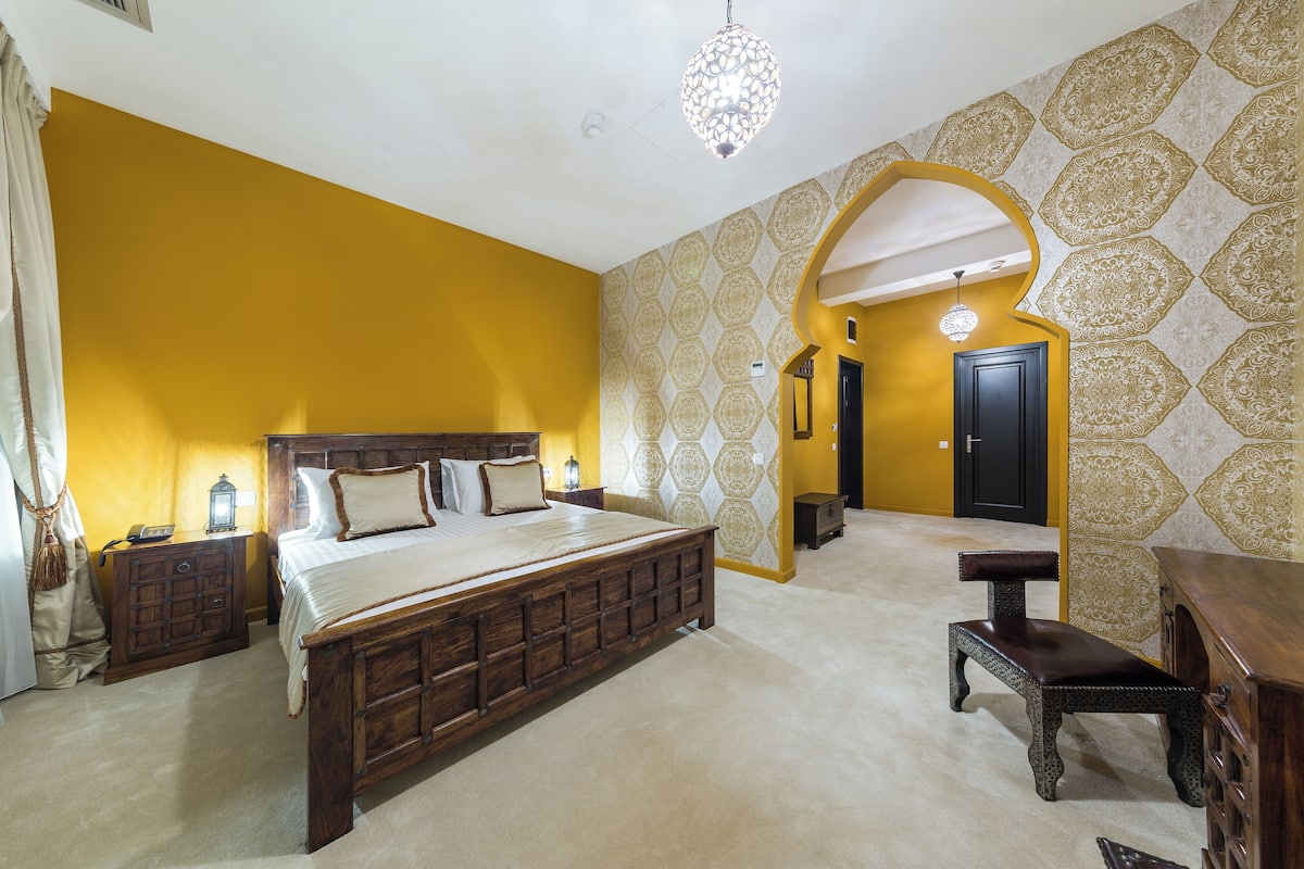A bedroom with a large bed, wooden nightstands, a bench, and a decorative wall pattern.