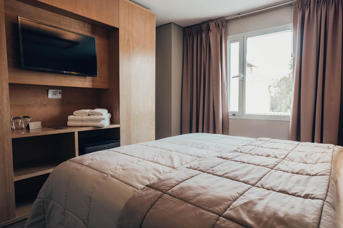 Une chambre d’hôtel avec un grand lit, un téléviseur à écran plat fixé au mur, une étagère avec des serviettes et une fenêtre avec des rideaux.