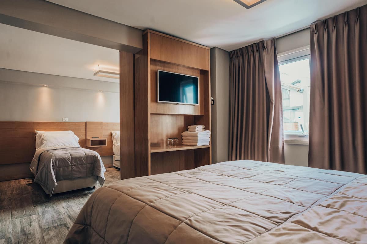 Une chambre d’hôtel avec un grand lit, un téléviseur fixé sur un panneau en bois, une table de chevet et une fenêtre avec des rideaux.