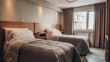 Habitación de hotel con dos camas, cabecero de madera, mesita de noche con lámpara, ventana con cortinas y radiador.