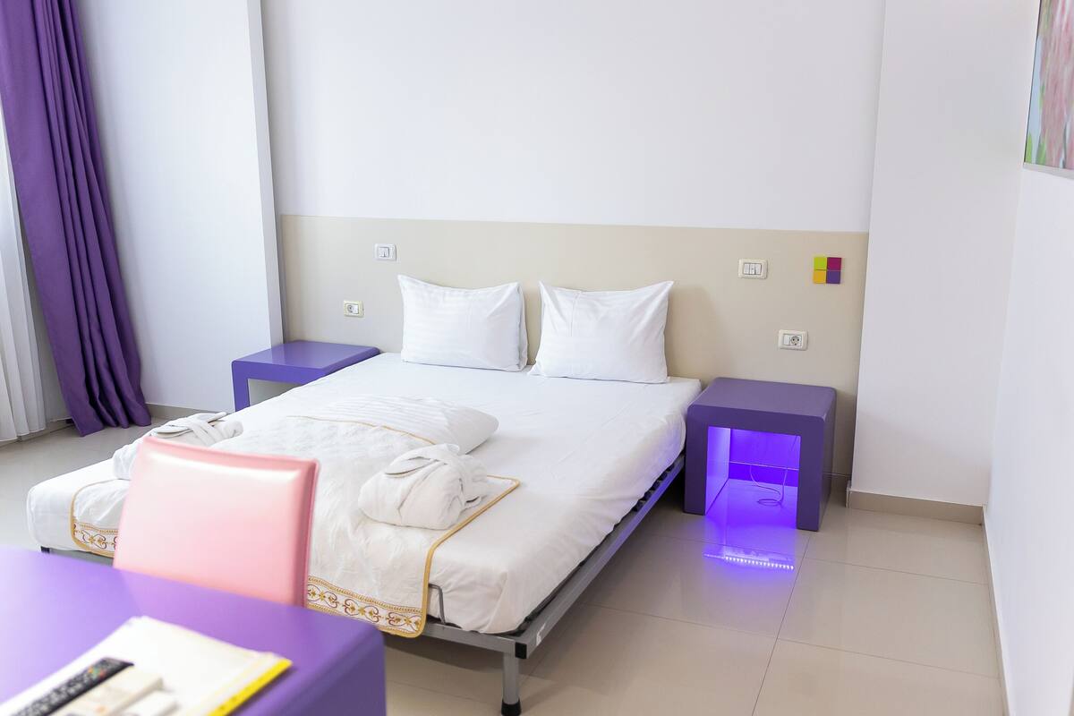 Un lit bien fait, avec du linge de lit blanc et des oreillers, une table de chevet avec une lampe et un fauteuil violet.