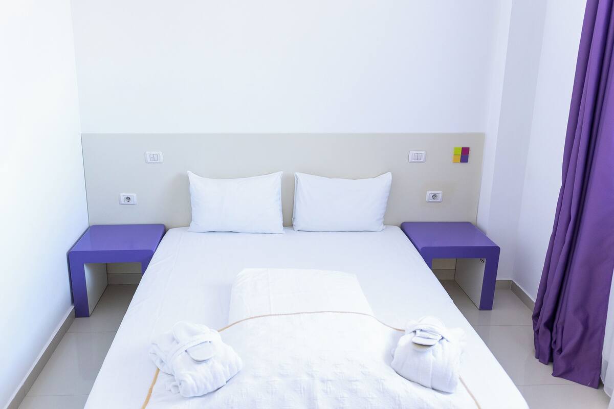 Un lit bien fait, avec du linge de lit blanc et deux oreillers, flanqué de deux tables de chevet violettes sur lesquelles sont posées de petites lampes.
