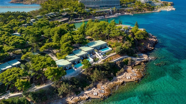Vista aérea de un complejo turístico costero con varios edificios, incluyendo un gran hotel y casas de huéspedes más pequeñas, rodeado de vegetación exuberante y aguas azules cristalinas.