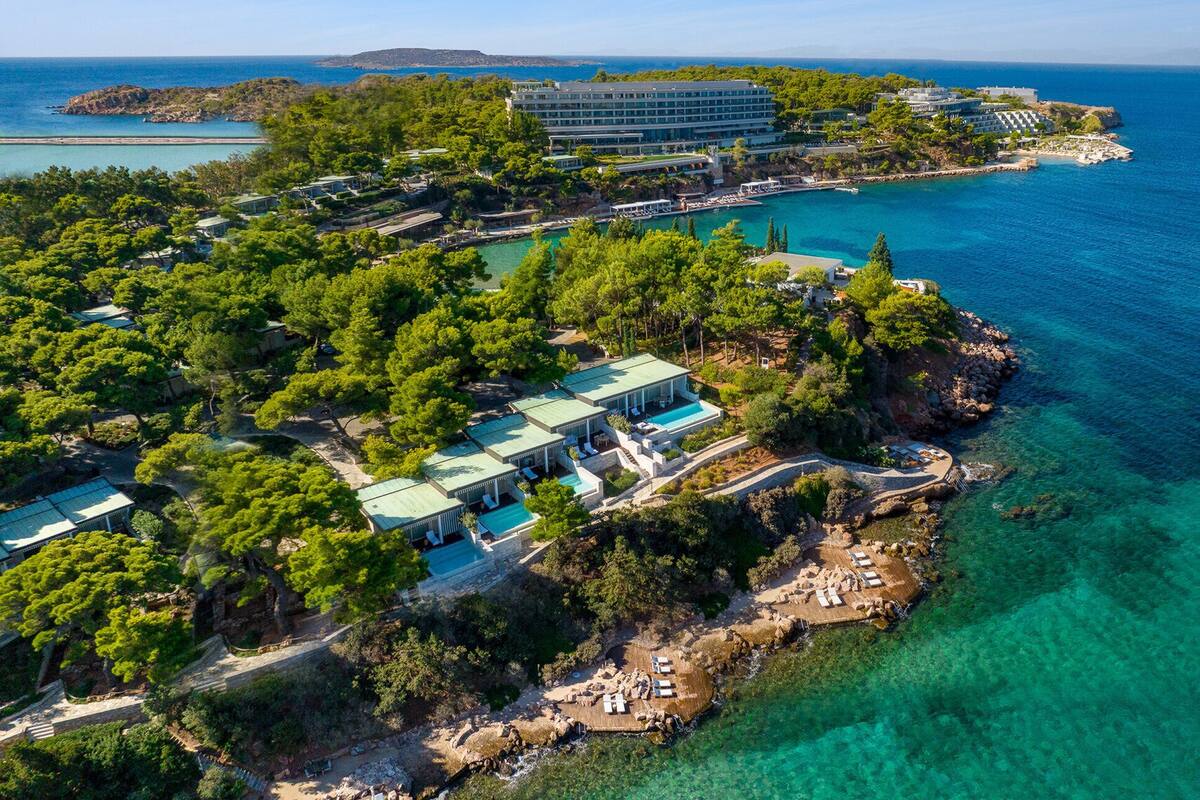 An aerial view of a coastal resort with multiple buildings, including a large hotel and smaller guest houses, surrounded by lush greenery and clear blue waters.