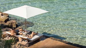Terraza de madera con sillones de descanso y una sombrilla con vistas a aguas azules y cristalinas.