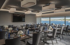 A hotel dining area with tables set for guests, large windows offering a view of the sea, and modern pendant lighting.