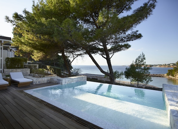 An infinity pool with a wooden deck, lounge chairs, and a view of the sea.