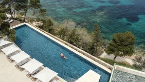 Piscina rectangular con una persona nadando, rodeada de sillas de descanso y sombrillas, con vistas a un mar azul y despejado.