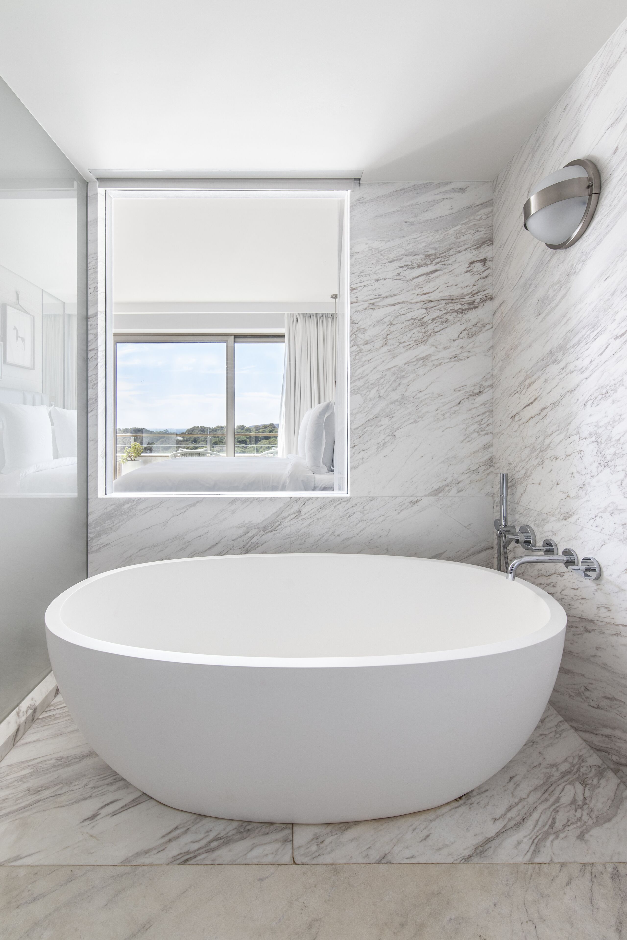 A modern bathroom with a large freestanding bathtub, marble walls, and a window offering a scenic view.