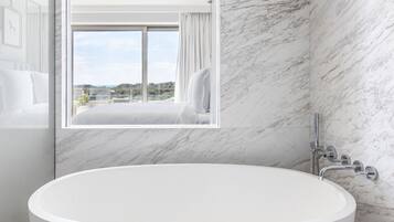 A modern bathroom with a large freestanding bathtub, marble walls, and a window offering a scenic view.