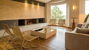 A modern living room with a fireplace, wooden flooring, and a large window offering a view of trees. - Langhetto Siena (Gramado)