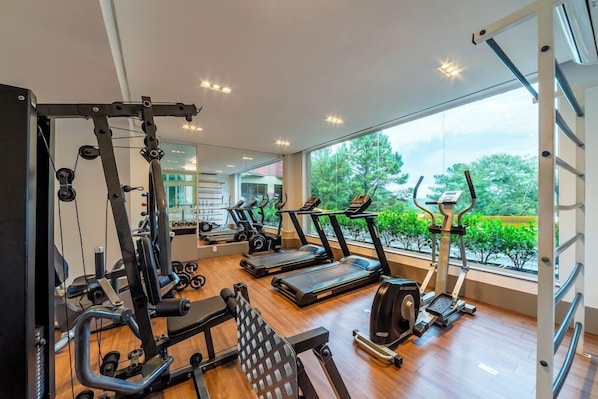 A gym with cardio machines, weight training equipment, and a large window with a view of greenery. - Langhetto Siena (Gramado)