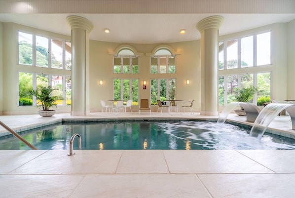 An indoor swimming pool with a waterfall feature, surrounded by columns and large windows offering views of greenery.