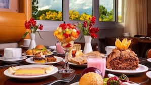 A breakfast buffet with various dishes, including a fruit salad, pastries, and a chocolate cake, set against a backdrop of a scenic view through large windows. - Langhetto Siena (Gramado)