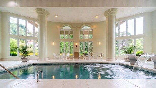 An indoor swimming pool with a waterfall feature, surrounded by columns and large windows offering views of greenery. - Langhetto Siena (Gramado)