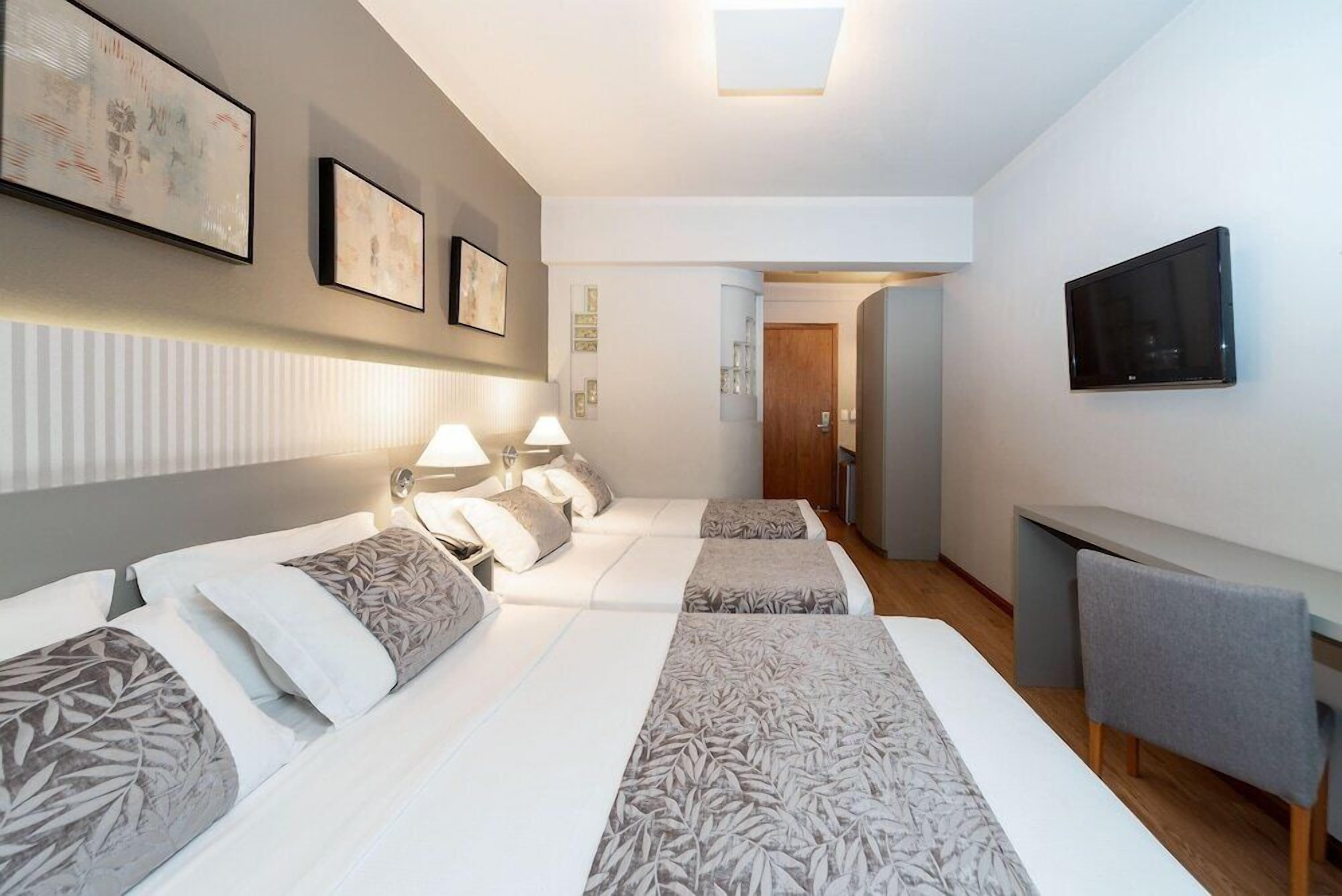 Quarto de hotel com cama, luminárias de cabeceira, televisão e uma pequena mesa.
