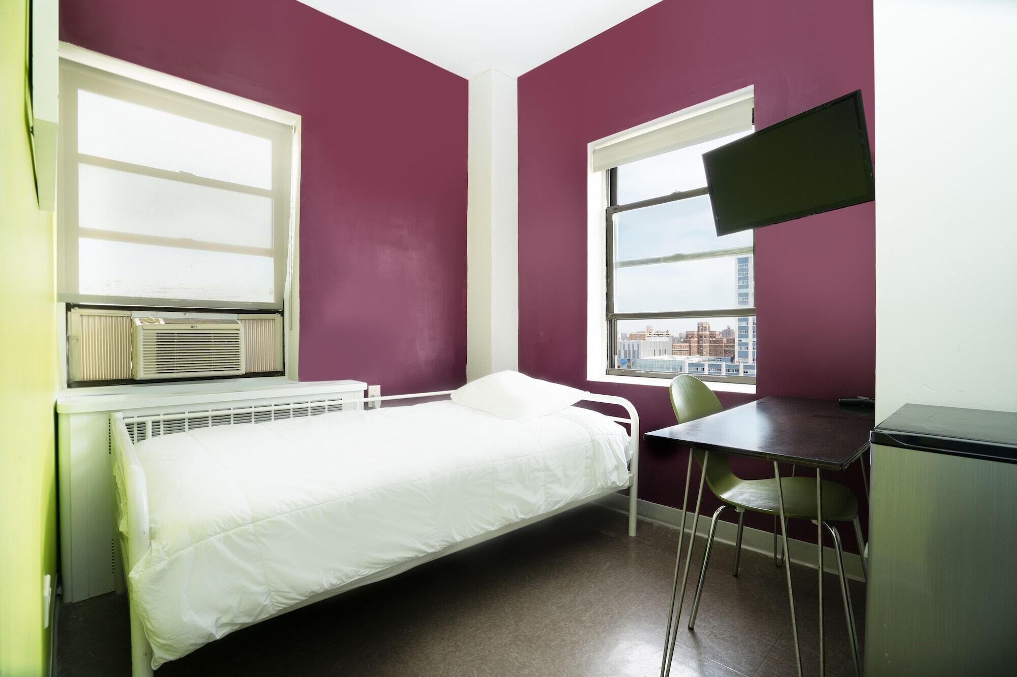 A small, single-bed room with a desk and chair, large windows, and an air conditioning unit.