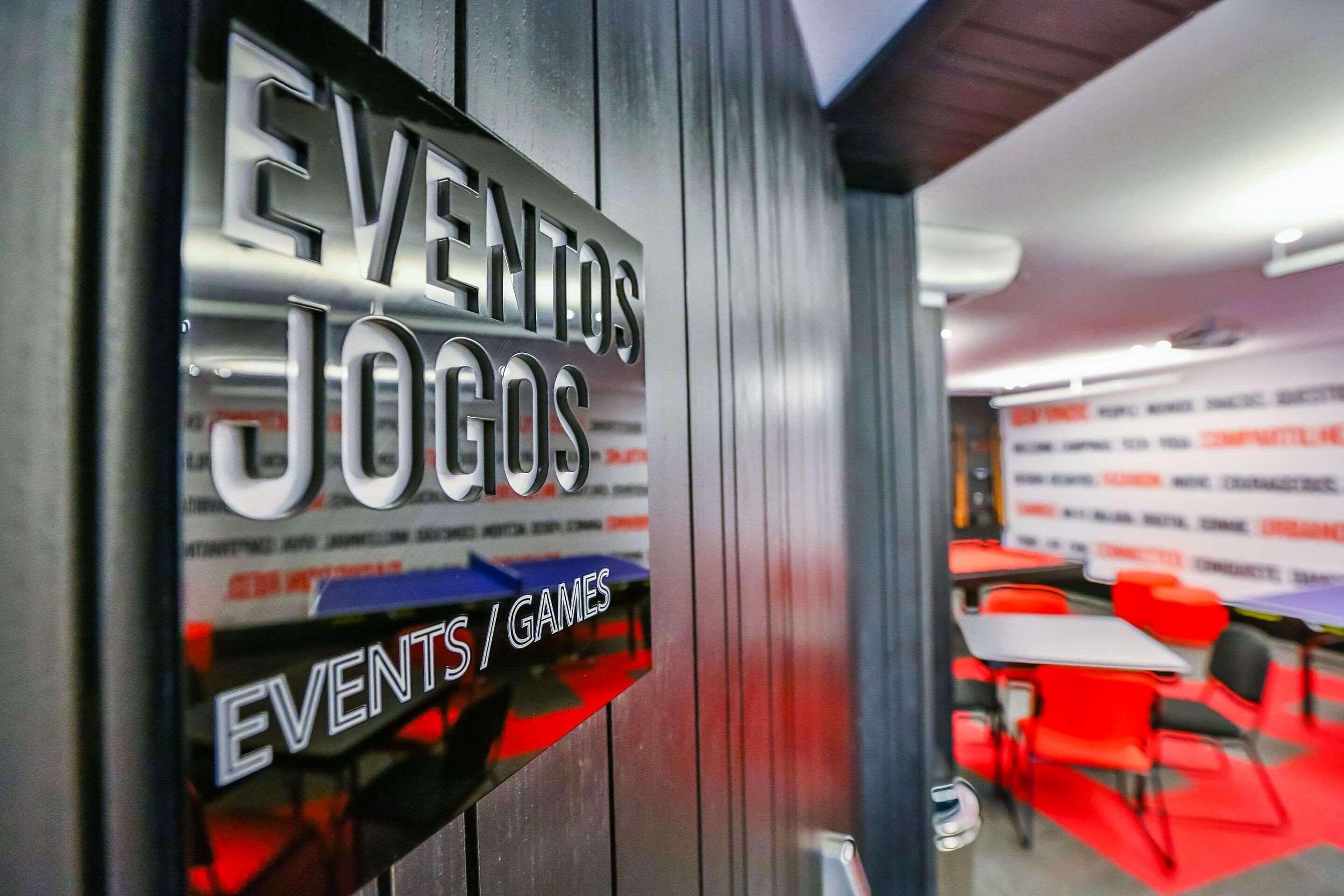 Señalización para 'EVENTOS JOGOS' en una sala con sillas y mesas rojas.