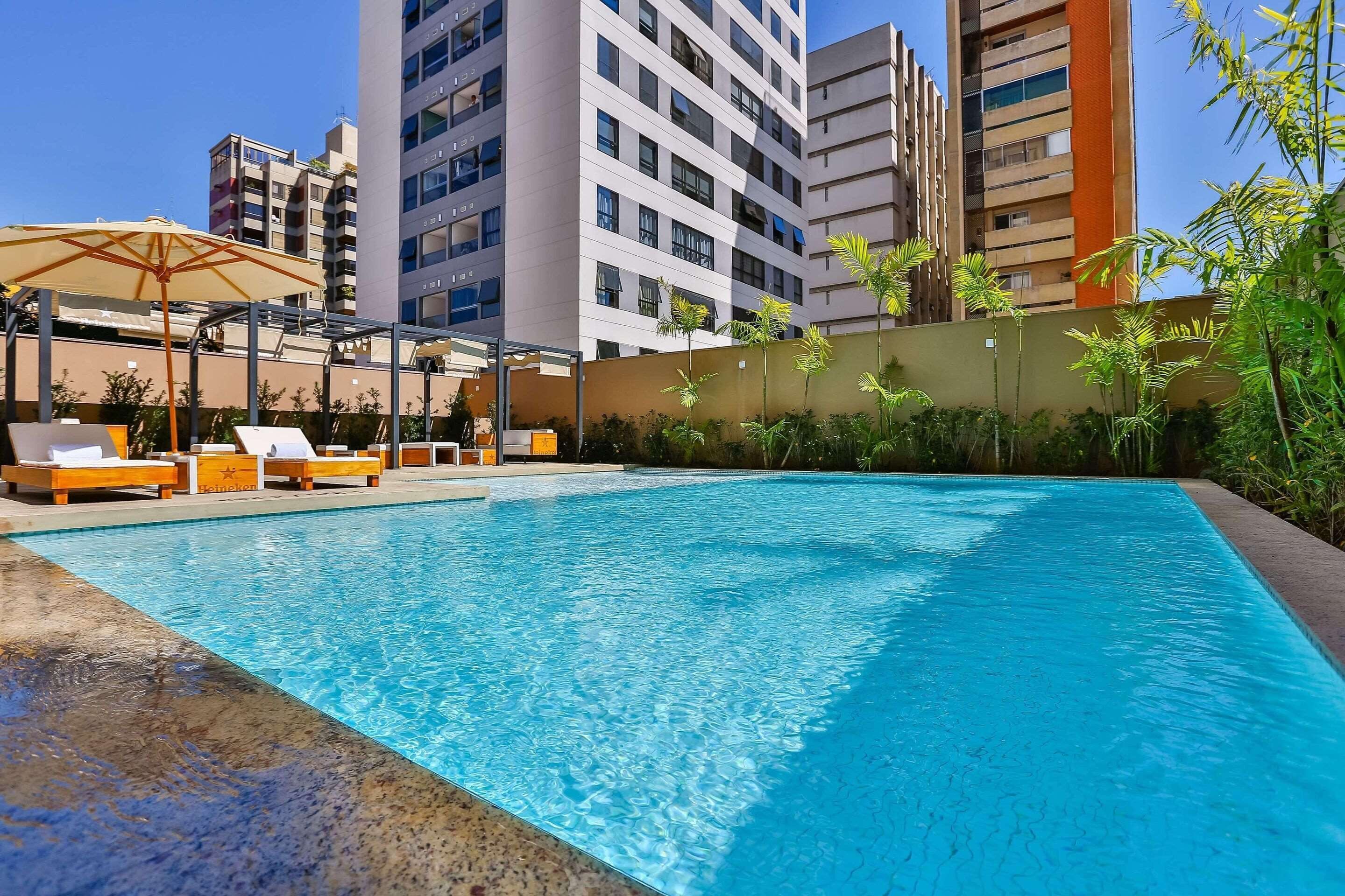 A swimming pool with lounge chairs and umbrellas, surrounded by tall buildings and greenery.