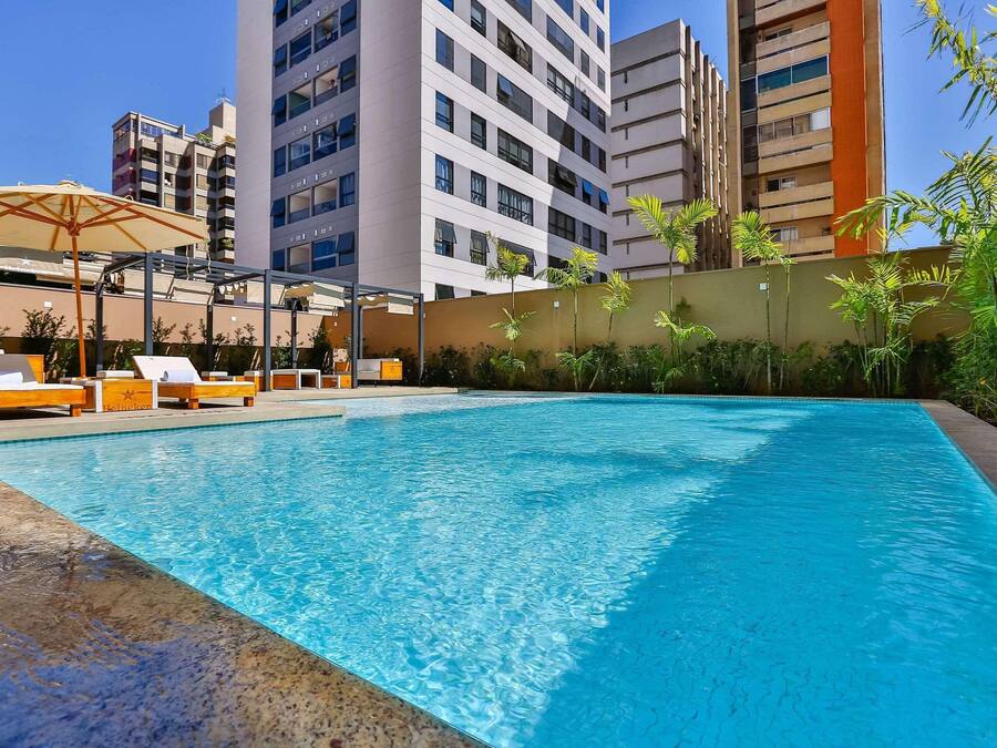 A swimming pool with lounge chairs and umbrellas, surrounded by tall buildings and greenery.
