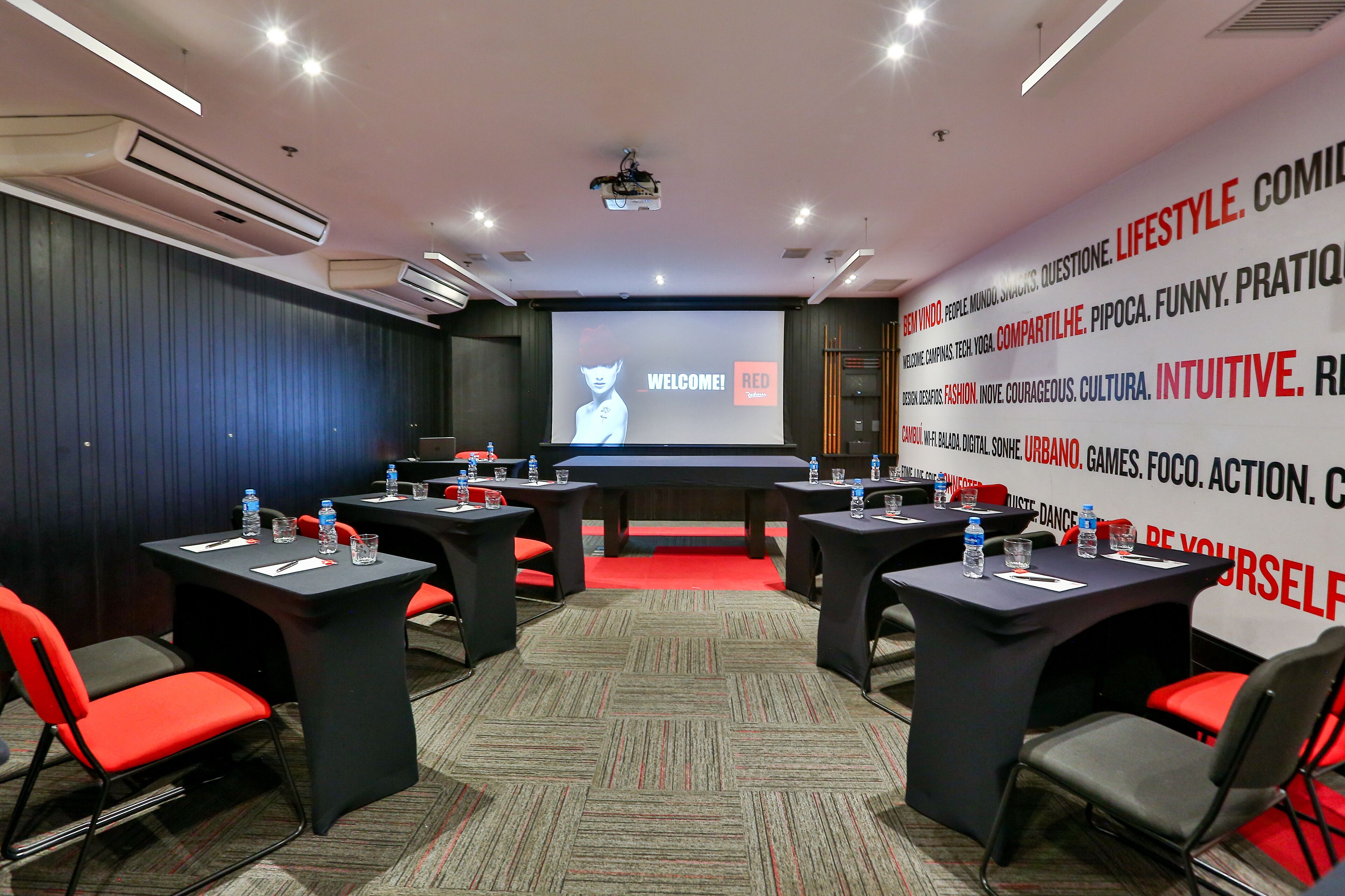 Un salón de conferencias con una pantalla grande mostrando un mensaje de bienvenida, varias mesas con sillas y una alfombra roja.