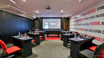 Un salón de conferencias con una pantalla grande mostrando un mensaje de bienvenida, varias mesas con sillas y una alfombra roja.