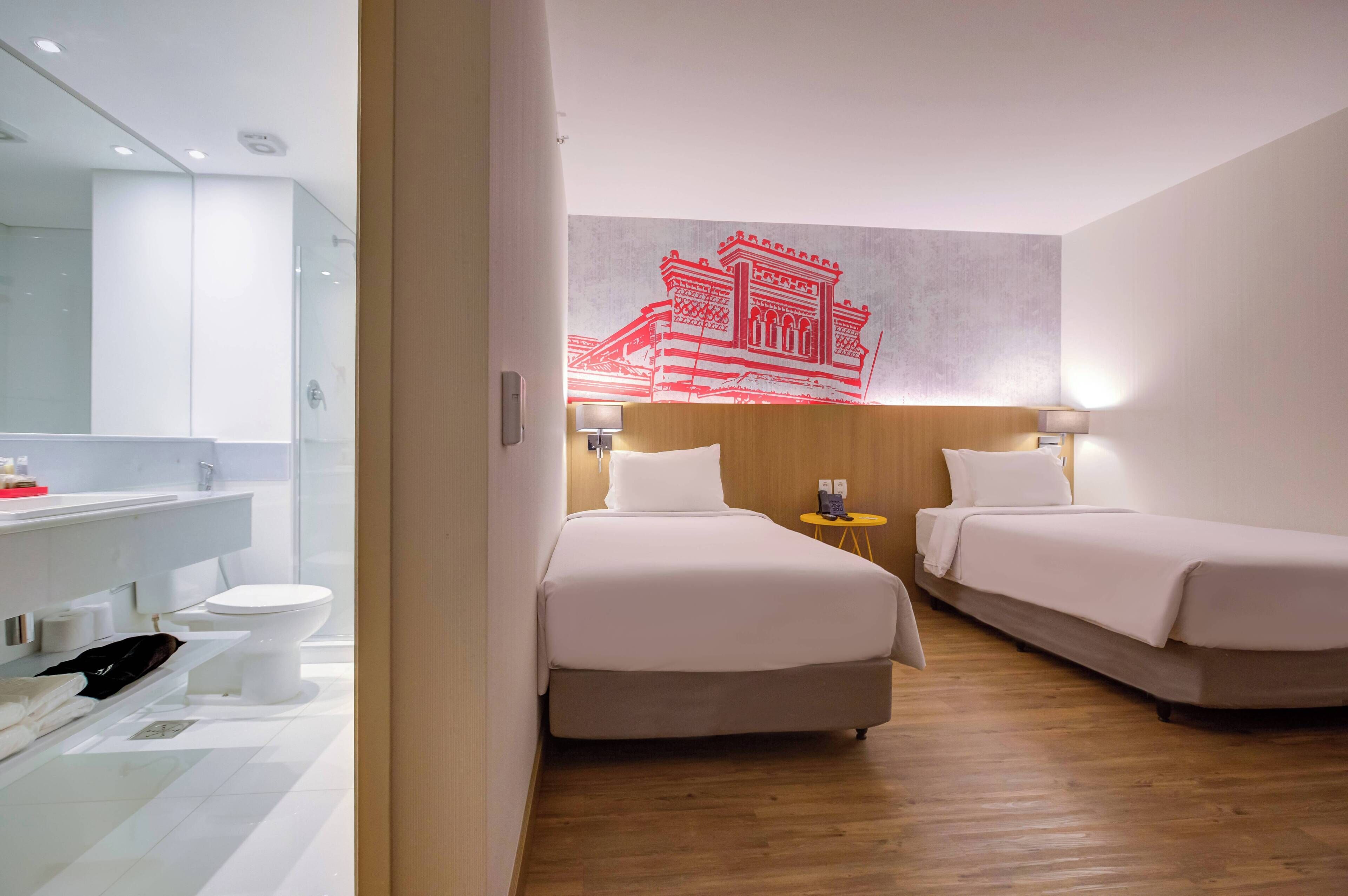 Habitación de hotel con suelo de madera, dos camas, baño con cabina de ducha de vidrio y una obra de arte colgada en la pared.