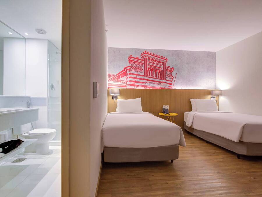 Habitación de hotel con suelo de madera, dos camas, baño con cabina de ducha de vidrio y una obra de arte colgada en la pared.