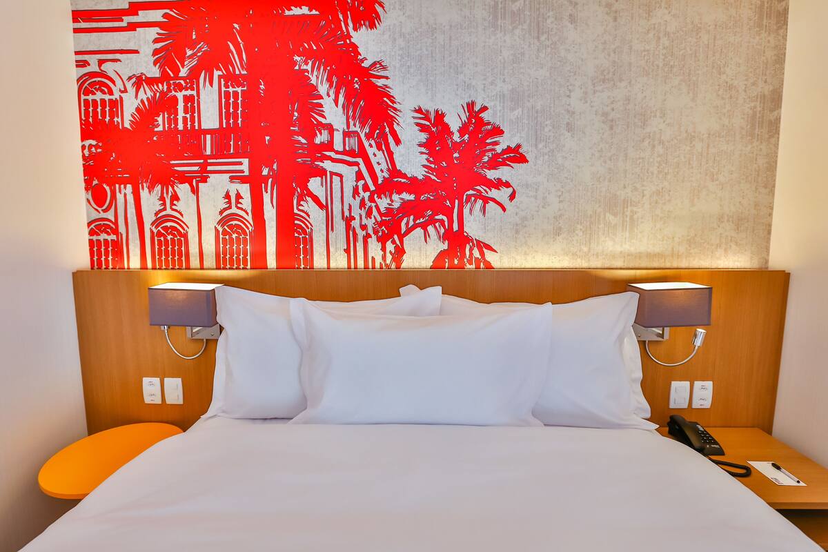 A hotel room with a bed, white pillows, a nightstand with a lamp, and a wall with a red abstract design.