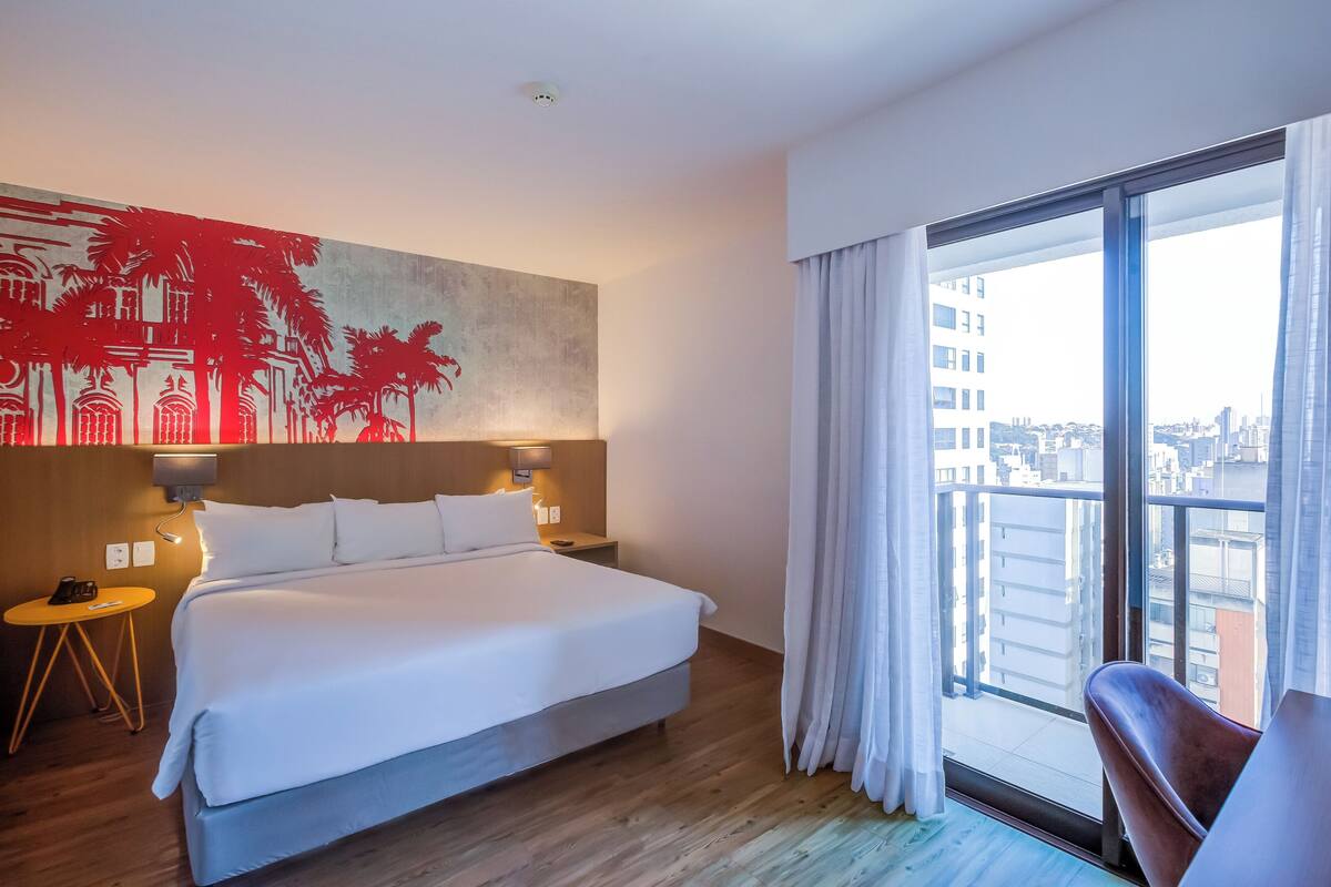 A hotel room with a large bed, a nightstand, a wall-mounted lamp, and a large window with curtains.