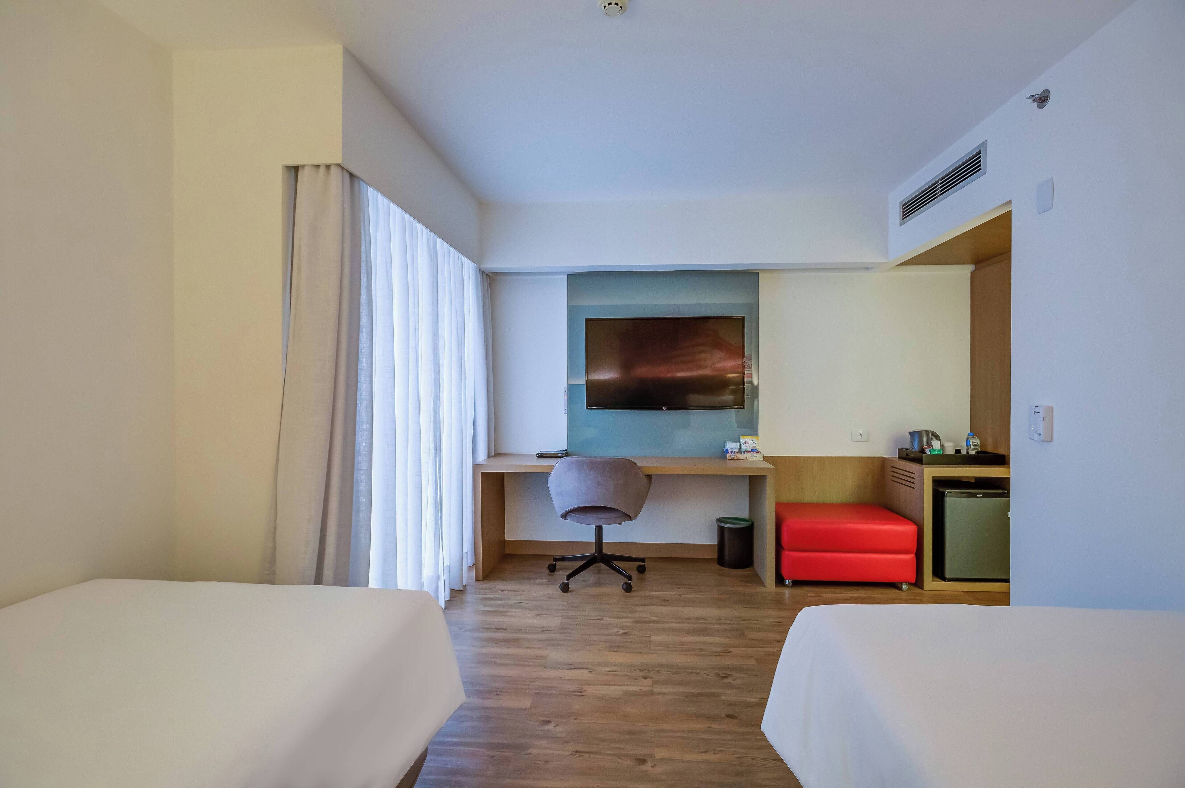 A hotel room with two beds, a desk with a chair, a TV, a small red sofa, and a mini-fridge.
