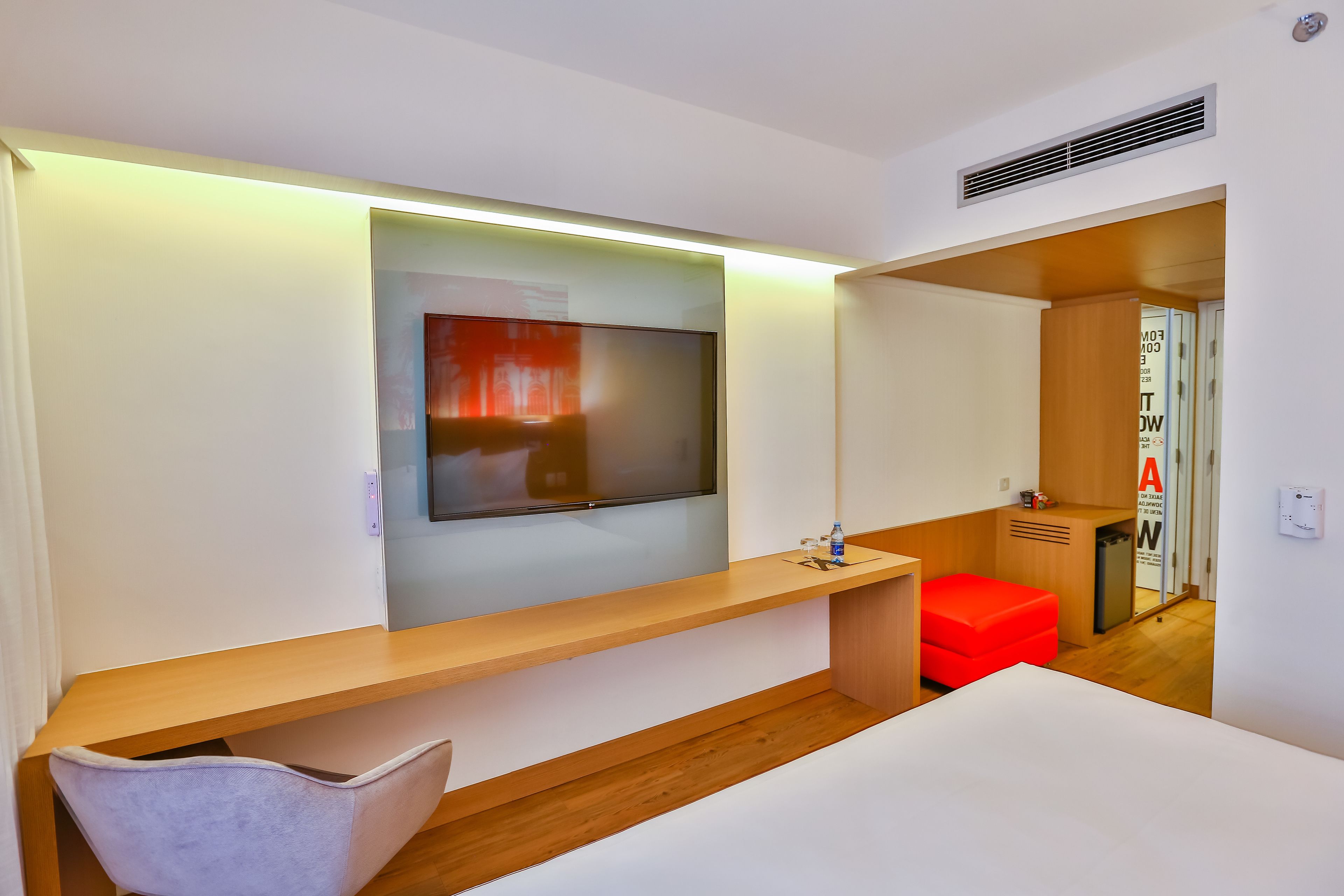 A modern hotel room with a large flat-screen TV mounted on the wall, a wooden desk, a red ottoman, and a white bed.