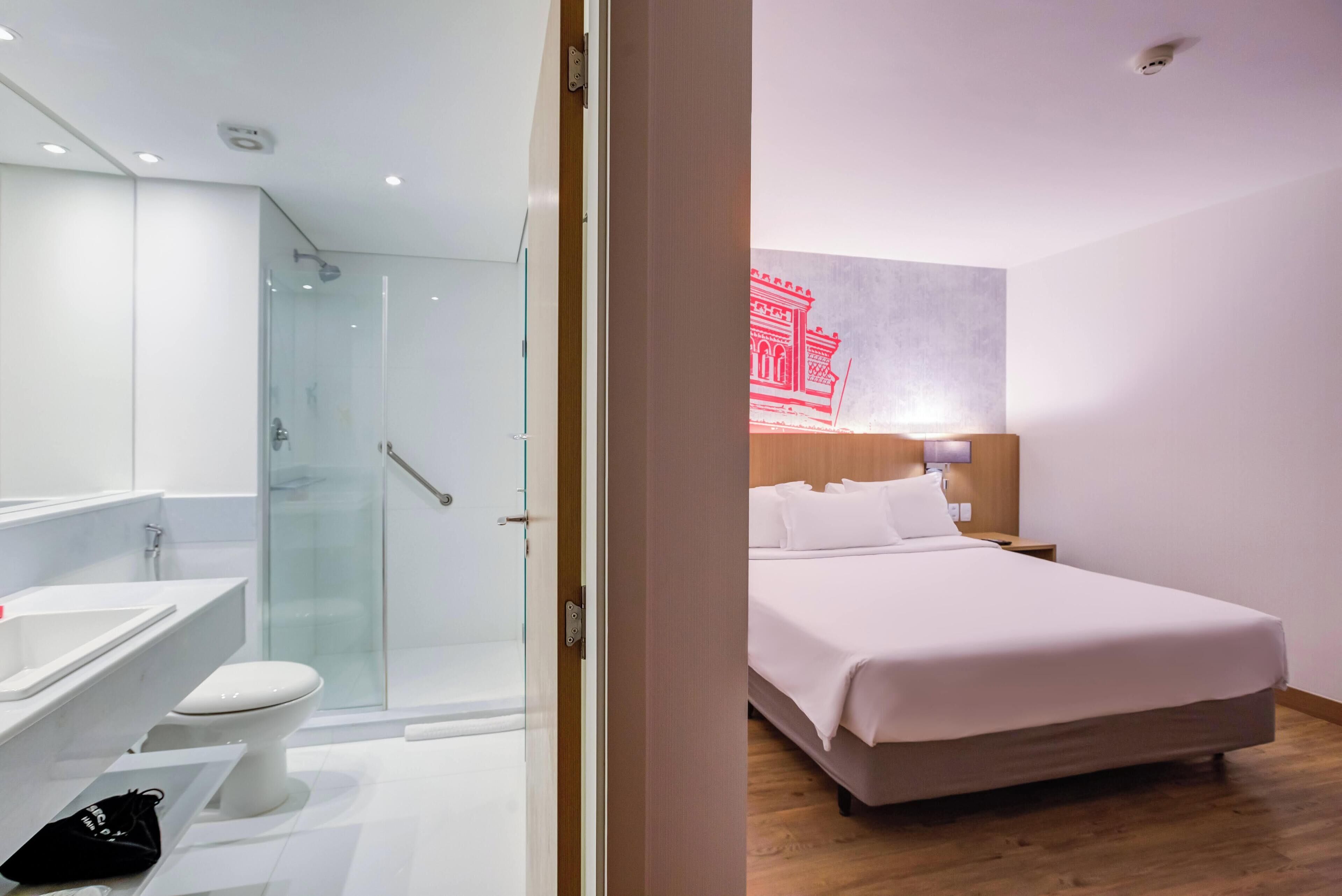 Ein modernes Hotelzimmer mit einem großen Bett, einer Duschkabine aus Glas, einem weißen Waschbecken und Holzboden.