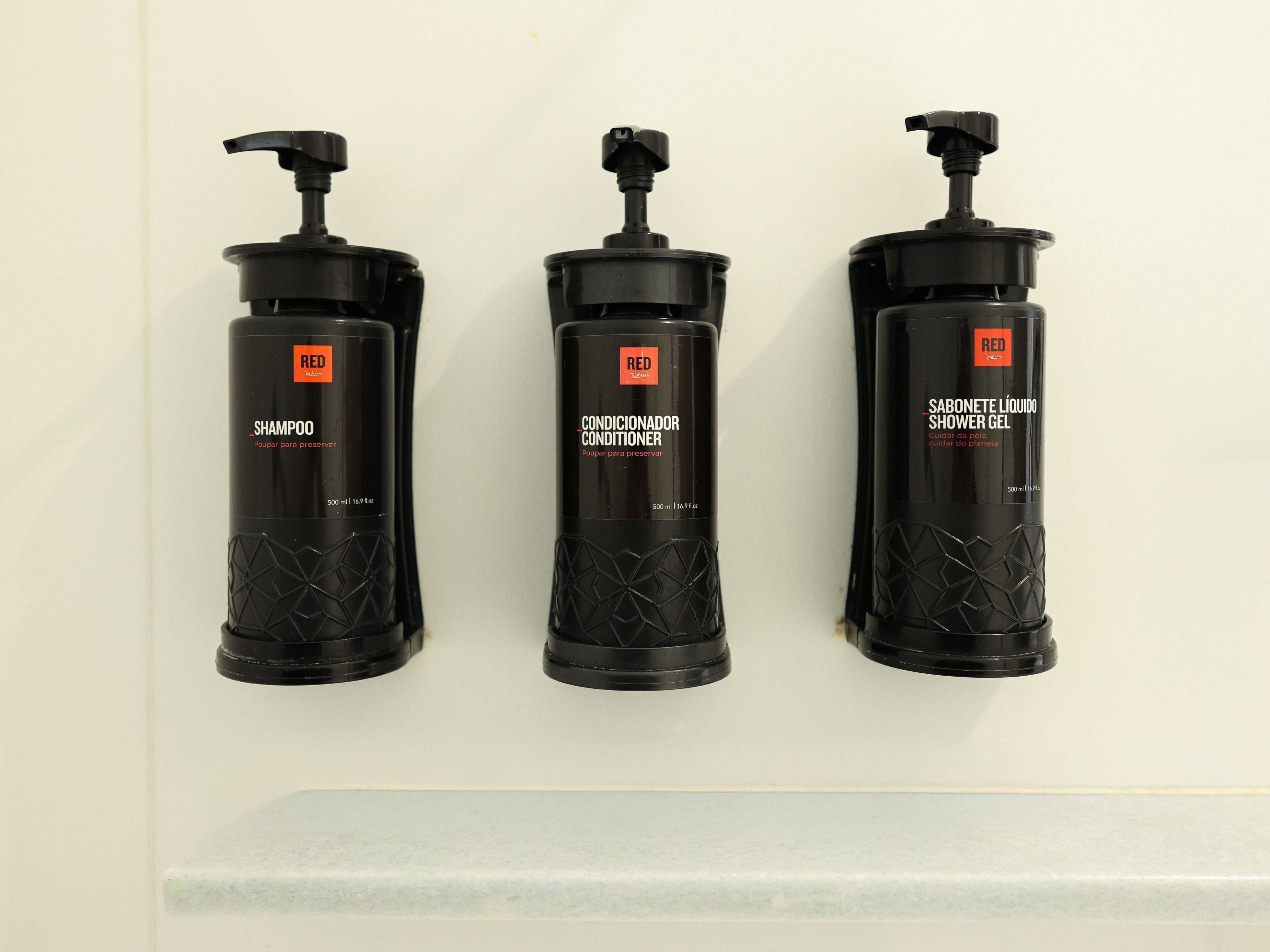 Three black bottles of Red shampoo, conditioner, and shower gel on a white wall.