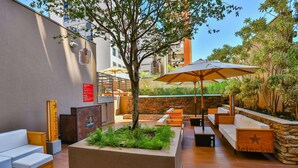 An outdoor seating area with wooden furniture, a central planter with greenery, and a large umbrella providing shade. - Radisson RED Campinas (Campinas)