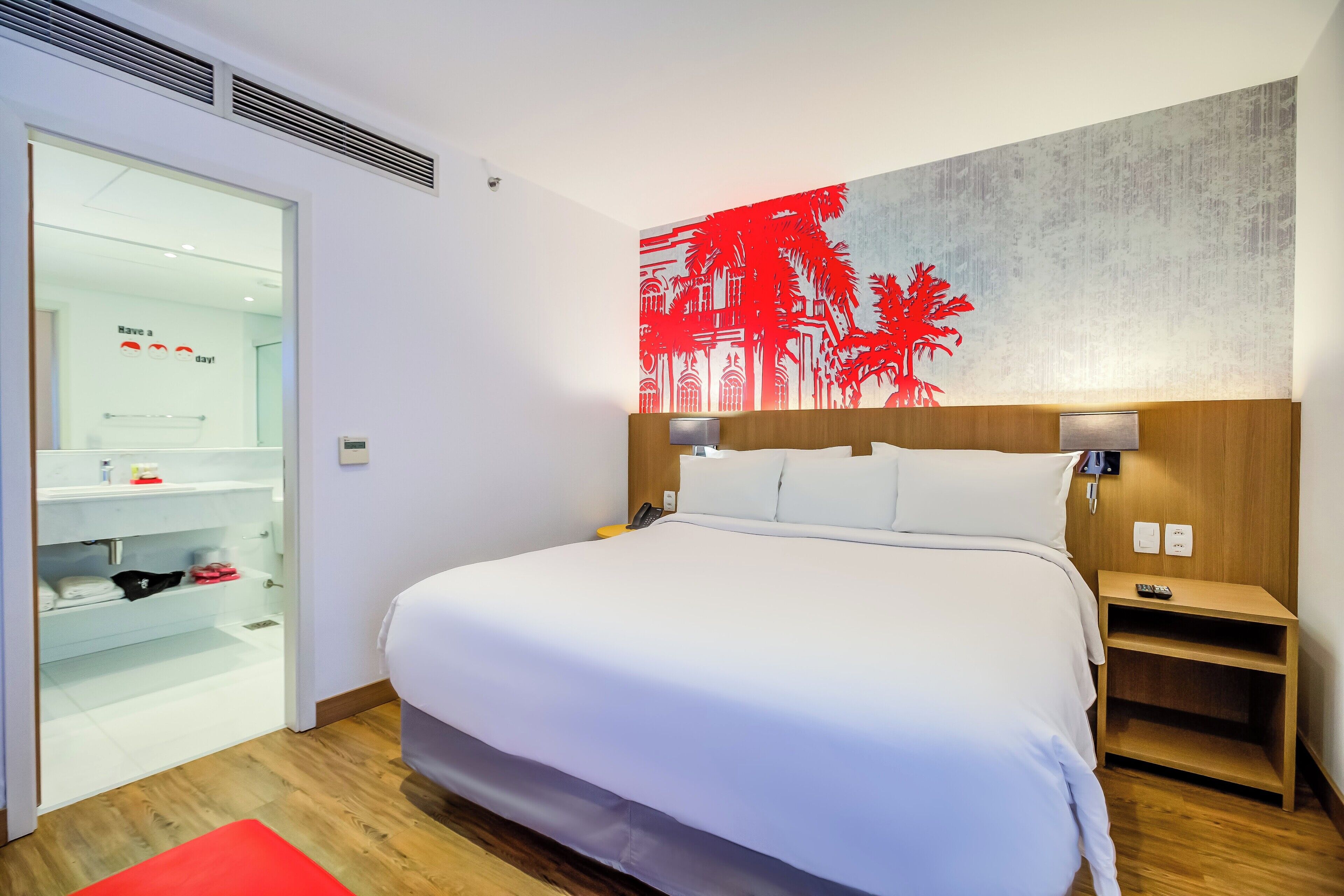 A modern hotel room with a large bed, a wooden headboard, a nightstand, a red rug, and a wall with a red palm tree and building design.