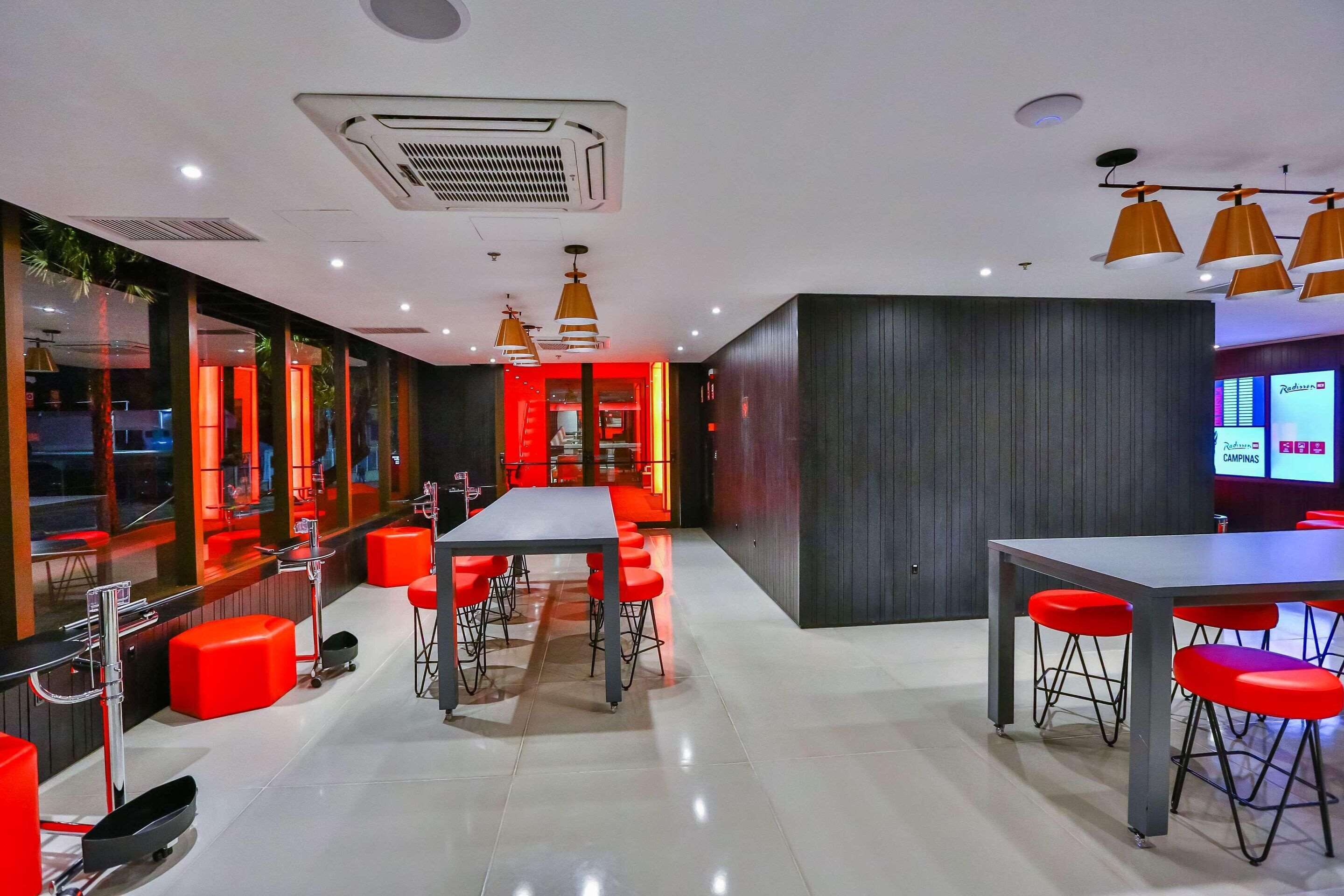 A modern restaurant with a long communal table, red bar stools, and a bright red accent wall.