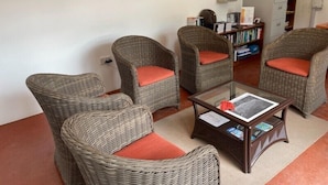 Una habitación con cuatro sillas de mimbre con cojines naranja, una mesa de centro de cristal y una estantería con libros y objetos decorativos.