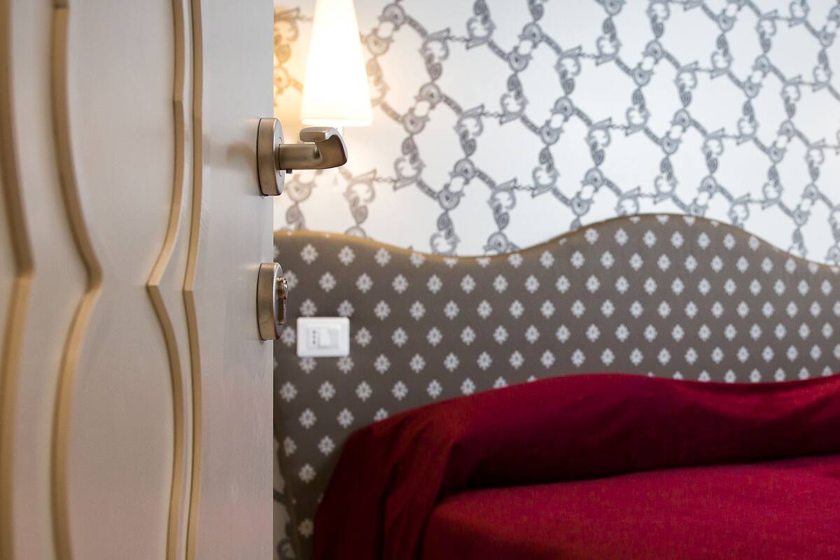 Una habitación de hotel con papel tapiz estampado, una cama con una manta roja y un enchufe de pared.