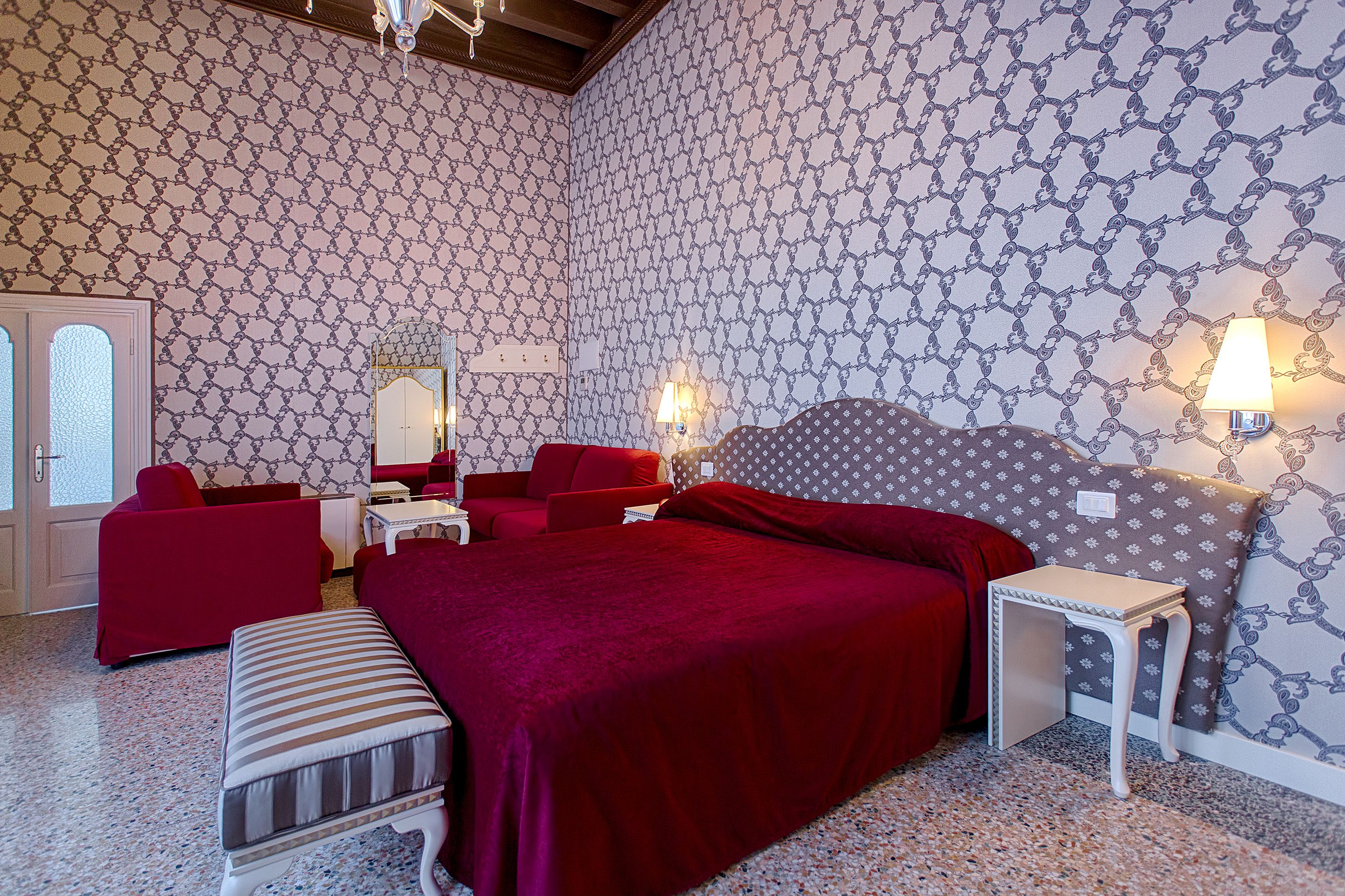 Un dormitorio con papel tapiz estampado, una cama grande con una manta roja, dos sillas rojas, una mesita y una mesita de noche.