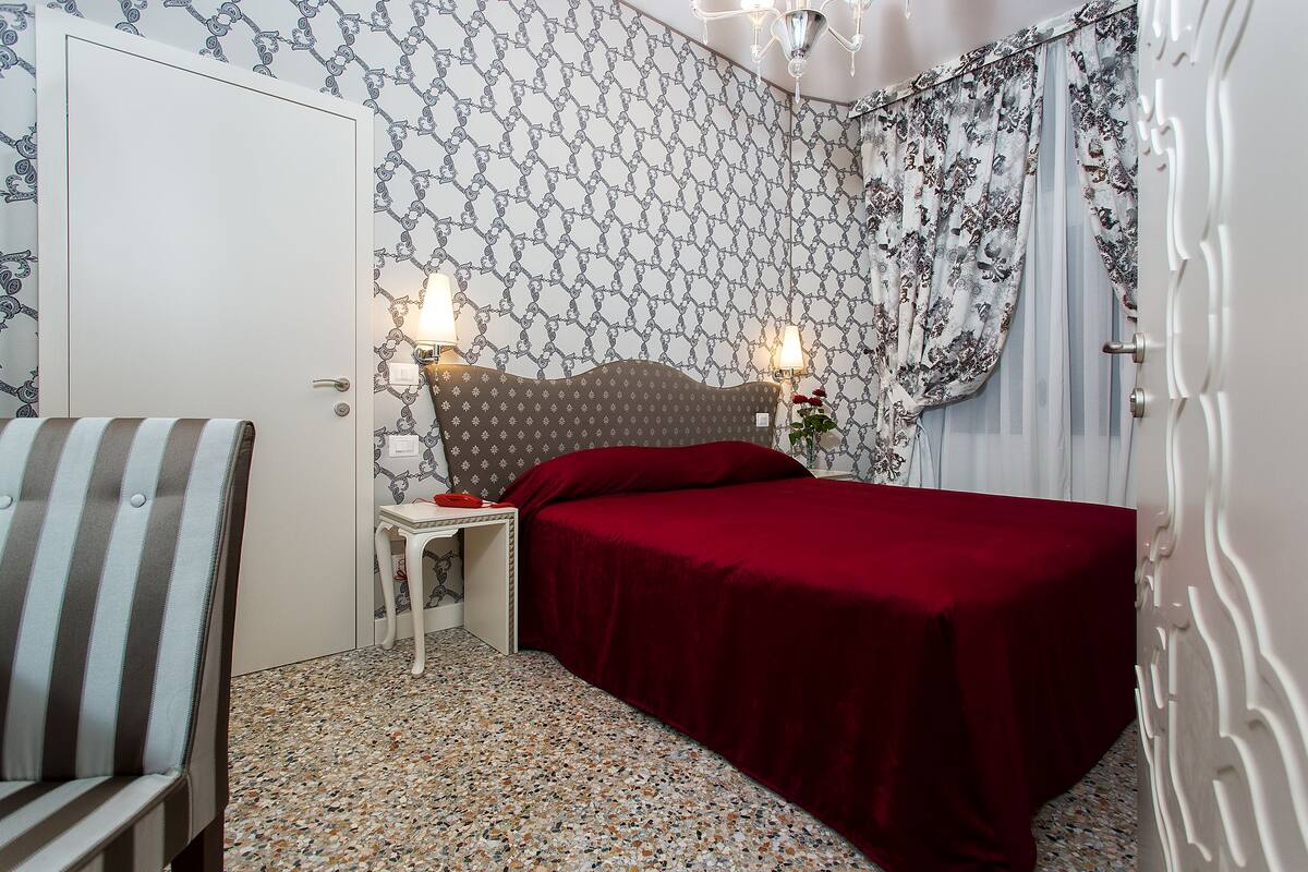 Un dormitorio con cama, mesita de noche, una silla y papel tapiz estampado.