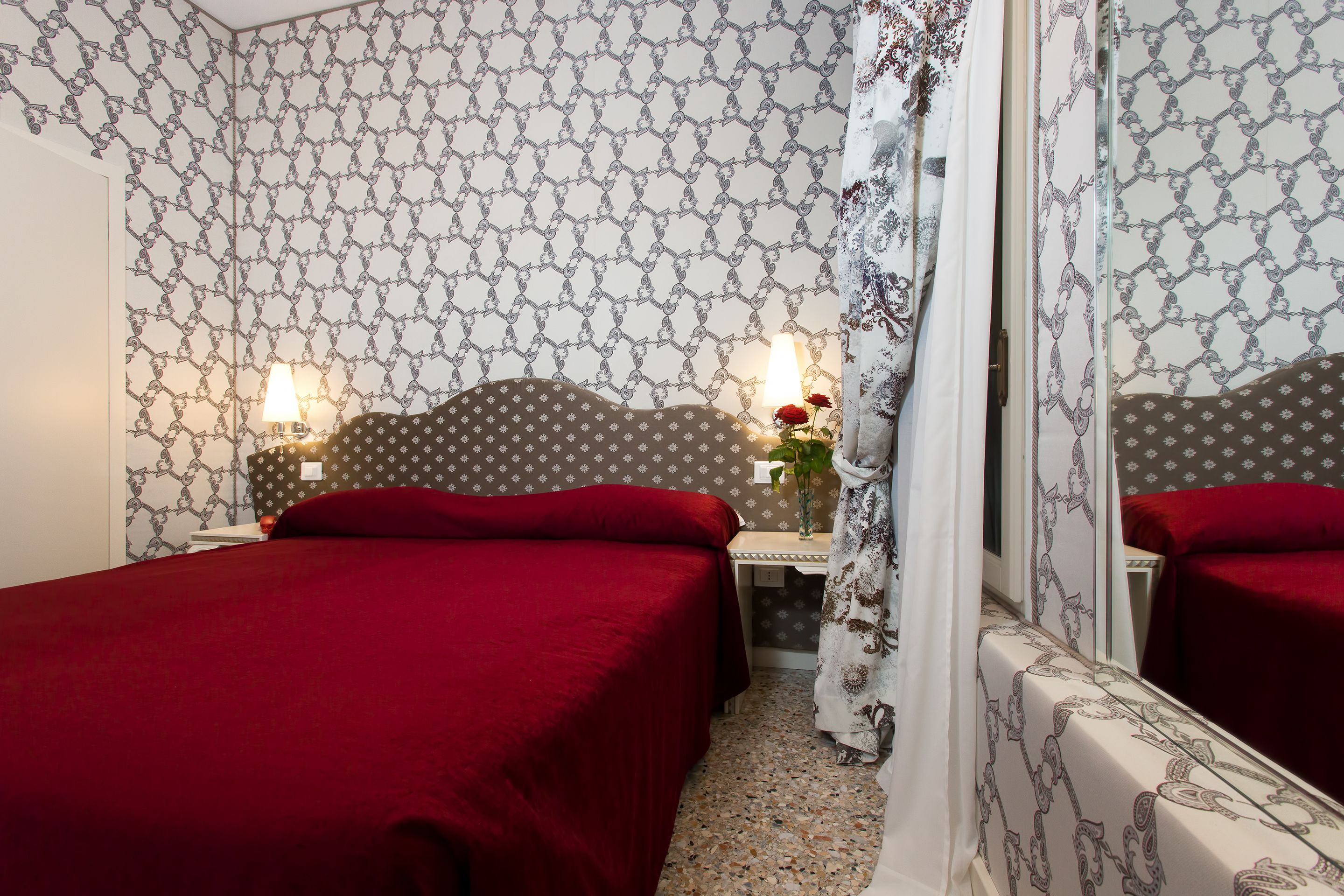 Una camera d'albergo con carta da parati decorata, un letto con copriletto rosso, un comodino con lampada e uno specchio che riflette la stanza.
