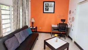 A room with a dark brown sofa, a small table with a vase, a desk with a chair, and an orange accent wall with a framed picture. - La Casa de Joy Airport Apartments (St. John's)