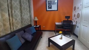 A living room with a black sofa, a coffee table, an orange accent wall, a framed picture, and a desk with a chair. - La Casa de Joy Airport Apartments (St. John's)