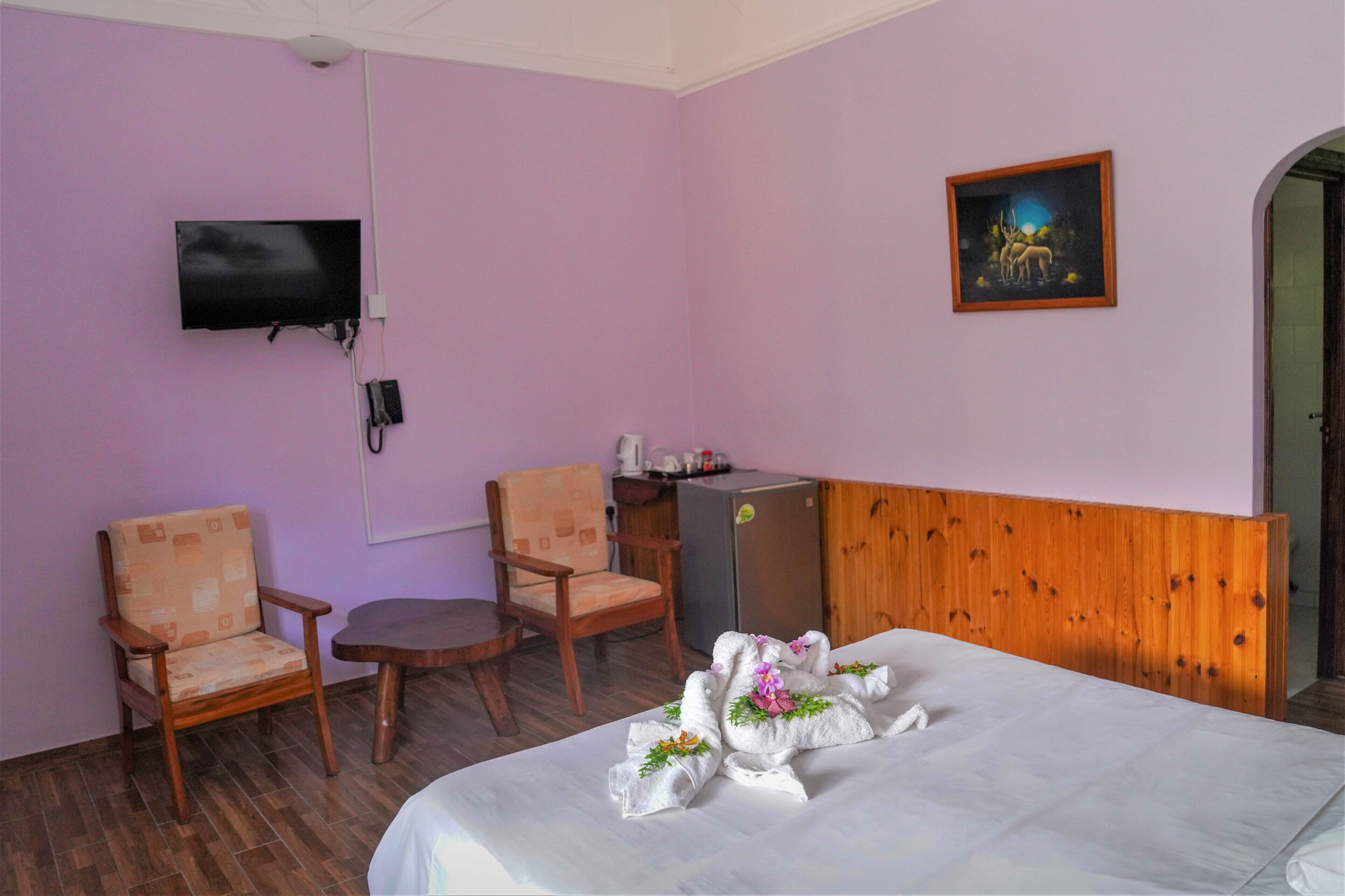 Une chambre d’hôtel avec un lit, deux chaises, une petite table, un téléviseur, un réfrigérateur et un tableau au mur.
